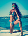 Woman in a blue latex bikini and sunglasses on a deck with water and sky in the background