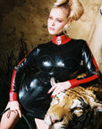 A person wearing a high-necked latex dress with red trim and contrast stripes, sitting next to a life-sized tiger mannequin.