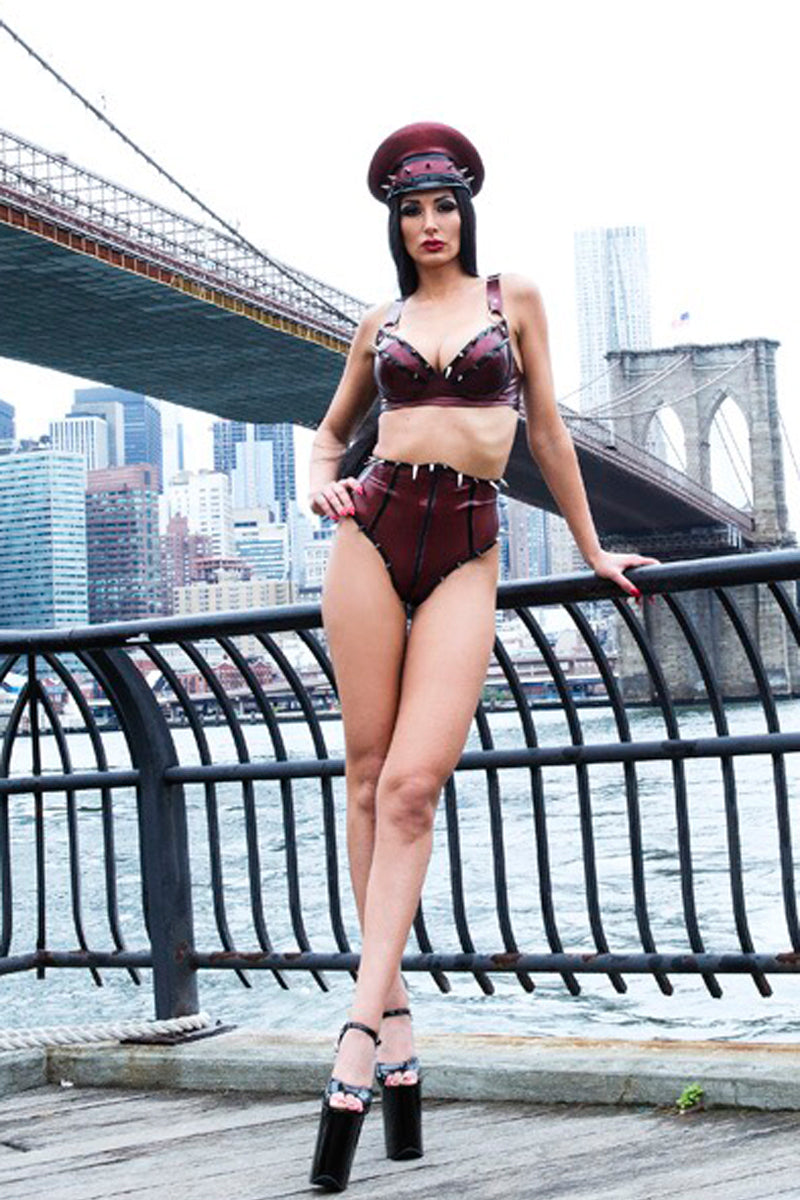 Person in burgundy latex lingerie and hat standing on a bridge with city skyline in the background