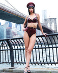 Person in burgundy latex lingerie and hat standing on a bridge with city skyline in the background